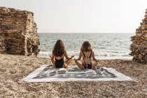 Back view of women in swimsuits sitting on blanket in lotus position while practicing yoga by stone construction — Stock Photo