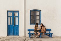 Щасливі молоді жінки в топах і шортах сидять на лавці і використовують мобільний телефон — стокове фото