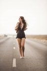 Atractiva joven mujer descalza caminando por un camino vacío, sosteniendo la mochila y mirando hacia otro lado - foto de stock