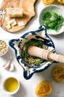 Top view of rustic pestle and mortar with smashed pine nuts and herbs next to cheese and raw pasta on kitchen table — Stock Photo