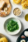 Top view of fresh green basil leaves on plates next to pine nuts and Parmesan cheese on white kitchen table — Stock Photo