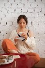 Mulher relaxada em roupas domésticas bebendo de caneca enquanto sentado à mesa da cozinha com comida ao lado da parede de tijolo branco — Fotografia de Stock