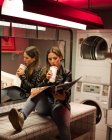 Mulher adolescente confiante em jaqueta de couro bebendo com palha de taça takeaway e revista de leitura enquanto espera pela lavanderia feita na lavanderia pública — Fotografia de Stock