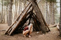 Side view of carefree blonde barefoot woman in bright leotard sitting on hunkers next to hut in pine forest — Stock Photo