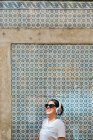 Mujer hermosa despreocupada en traje casual y auriculares de pie en la pared de mosaico azul del edificio en la calle de la ciudad - foto de stock