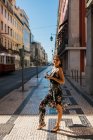 Confident beautiful woman in summer dress holding a photo camera while standing on scenic sunny city street in Lisbon, Portugal — Stock Photo