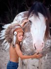 Tranquil child with closed eyes wearing traditional Indian war bonnet, bonding with horse stallion on blurred background — Stock Photo
