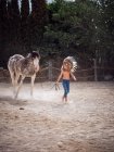 Calm kid wearing feather Indian war bonnet and walking shirtless on sandy farm leading horse behind — Stock Photo