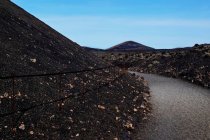 Schmale dunkle Straße in vulkanischen Mineralien, die zu felsigen Hügeln unter blauem Himmel auf Lanzarote, Kanarische Inseln, Spanien führt — Stockfoto