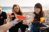 Mulher sorridente dá um pedaço de melancia para sua amiga com sua amiga que bebe suco de laranja na praia — Fotografia de Stock