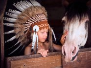 Очарованный ребенок в настоящей индийской шляпе из перьев опирается на деревянный забор в конюшне с лошадью на ранчо и смотрит в сторону — стоковое фото