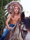 Chico serio en sombrero de plumas indio auténtico montar a caballo en el parque - foto de stock