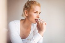 Young woman with blond hair and in bodysuit looking away while sitting on soft bed against white wall at home — Stock Photo
