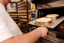 Взрослый мужчина в форме во время работы в пекарне кладет поднос с сырыми тортами в горячую печь — стоковое фото