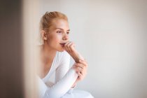 Young woman with blond hair and in bodysuit looking away while sitting on soft bed against white wall at home — Stock Photo