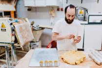 Homem barbudo em t-shirt branca colocando massa fresca em copos enquanto faz pastelaria na cozinha da padaria — Fotografia de Stock