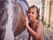 Happy shirtless kid with curly wet hair embracing horse side — Stock Photo