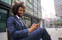 Empleado afroamericano feliz sentado en el banco y sonriendo mientras navega por las redes sociales usando un teléfono inteligente - foto de stock