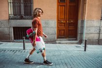 Side view of handsome sportive man holding longboard while walking on city summer city street — Stock Photo