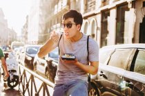 Bonito ásia homem no sol óculos comer takeaway alimentos com pauzinhos enquanto em pé por comida caminhão no ensolarado rua — Fotografia de Stock