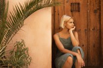 Junge Frau schaut weg, während sie neben Holztür und Pflanzen im Hof sitzt — Stockfoto