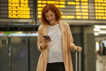 Счастливая рыжая девушка с чемоданом улыбается и разговаривает по мобильному телефону на вокзале — стоковое фото