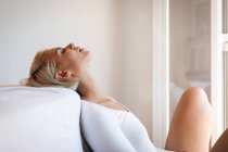 Vista lateral da bela mulher loira fechando os olhos e inclinando-se no colchão confortável contra a parede branca em casa — Fotografia de Stock