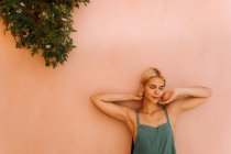 Bella giovane femmina con i capelli biondi corti chiudendo gli occhi e appoggiandosi alla parete mentre in piedi su sfondo rosa sfocato — Foto stock