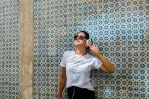 Bela mulher em roupa casual e fones de ouvido com as mãos para cima ao lado parede de mosaico azul de construção na rua da cidade — Fotografia de Stock