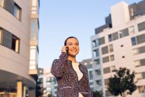 Mujer emprendedora alegre sonriendo y mirando hacia otro lado mientras habla en el teléfono inteligente - foto de stock