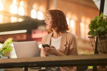 Весела молода жінка працює на ноутбуці в кафе і переглядає телефон — стокове фото