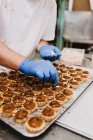 Невпізнаваний чоловік в латексних рукавичках кладе горіхи на поверх солодких дрібних тортів, працюючи в хлібобулочних виробах — стокове фото