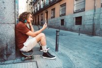 Вид сбоку случайного человека в наушниках, просматривающего смартфон, сидя на лонгборде у здания на улице — стоковое фото