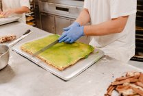 Человек в форме и перчатках с помощью ножа разрезать сладкий свежий торт на столе в пекарне — стоковое фото