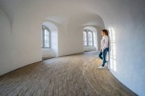 Side view of calm woman in jeans leaning on white wall at minimalistic hall with arched windows — Stock Photo