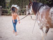 Back view of kid wearing feather Indian war bonnet and walking shirtless on sandy farm, leading horse behind — Stock Photo