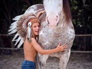 Tranquil child looking at camera wearing traditional Indian war bonnet, bonding with horse stallion on blurred background — Stock Photo