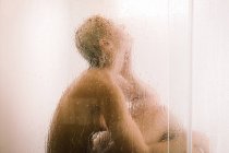 Vista lateral de la joven hembra tomando ducha detrás de la partición transparente húmeda en el baño - foto de stock