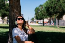 Мирна жінка в сонцезахисних окулярах і навушниках слухає музику, сидячи на траві в парку — стокове фото