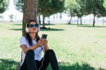 Мирна жінка в сонцезахисних окулярах і навушниках, використовуючи смартфон і слухаючи музику, сидячи на траві в парку — стокове фото