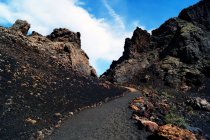 Estrada escura estreita em minerais vulcânicos subindo a colinas rochosas sob céu azul em Lanzarote, Ilhas Canárias, Espanha — Fotografia de Stock