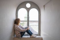 Vista lateral de la mujer moderna soñando sentado en el alféizar y mirando por la ventana arqueada - foto de stock
