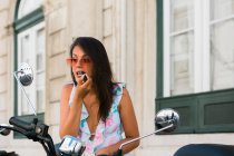 Carefree beautiful woman in sunglasses putting on bright lipstick while looking at motorbike mirror on street — Stock Photo