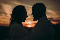 Vista lateral silueta de la pareja de cuidados abrazos y mirarse unos a otros mientras que están en el campo en el cielo oscuro rojo puesta de sol. - foto de stock