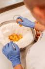 From above unrecognizable confectioner in latex gloves breaking fresh chicken eggs into bowl with wheat flour while preparing pastry in kitchen — Stock Photo