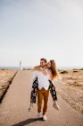Loving man giving piggyback tide to woman while walking on road from lighthouse and sea in summer and looking away — Stock Photo