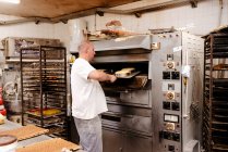 Uomo in uniforme mettere vassoio con torte crude in forno caldo mentre si lavora in panetteria — Foto stock