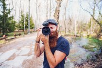 Современный бородатый рыжий мужчина в кепке фотографируется с камерой, стоя на дороге в окружении лиственных деревьев в красочных лесах в солнечный осенний день — стоковое фото