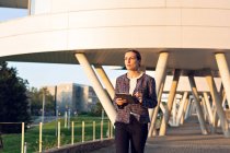Grave donna d'affari in elegante giacca tablet di navigazione e guardando lontano mentre in piedi sulla strada della città nella giornata di sole — Foto stock