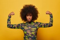 African American empowered woman in camouflage shirt showing biceps on yellow background looking at camera — Stock Photo